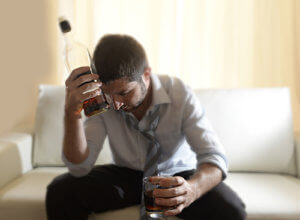 Young man on sofa holding bottle to head needing alcohol abuse treatment.